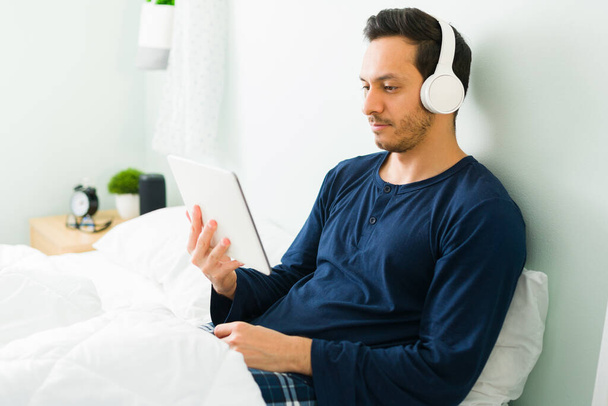 Young man resting in his comfy bed and enjoying a video on his tablet. Latin man relaxing in his bedroom while watching a movie with headphones  - Foto, Imagem