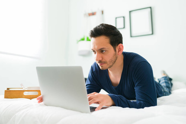 Attractive young man in pajamas lying on his stomach in bed while typing on a laptop next to a tray with breakfast - Fotó, kép