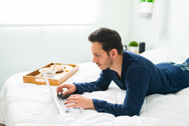 Расслабленный человек печатает на ноутбуке и делает покупки в интернете, лежа утром в постели. Испанец наслаждается завтраком в спальне - Фото, изображение