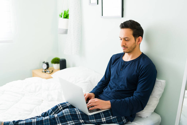 Atraktivní muž, který píše na notebook a pracuje, zatímco sedí na posteli. Hispánec muž v pyžamu dělá on-line nakupování během volného času - Fotografie, Obrázek