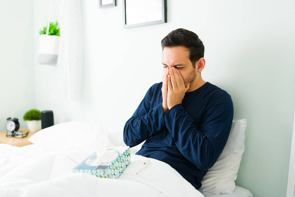 Homem latino doente vestindo pijama e soprando o nariz com um tecido por causa da gripe enquanto descansa na cama  - Foto, Imagem
