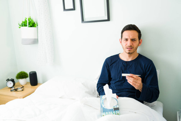 Πορτραίτο ενός άρρωστου άντρα που κάνει οπτική επαφή κρατώντας ένα θερμόμετρο για τον πυρετό του. Αναστατωμένος άνθρωπος με γρίπη αισθάνεται άσχημα ενώ ξεκουράζεται στο κρεβάτι - Φωτογραφία, εικόνα