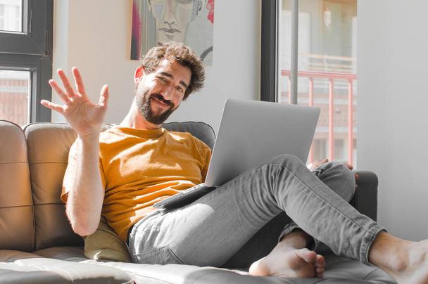 young bearded man smiling happily and cheerfully, waving hand, welcoming and greeting you, or saying goodbye with a laptop on a couch laptop concept - Fotografie, Obrázek