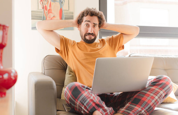 молодий бородатий чоловік відчуває стрес, тривогу, тривогу або наляканий, з руками на голові, панікуючи помилково і сидячи з ноутбуком
 - Фото, зображення