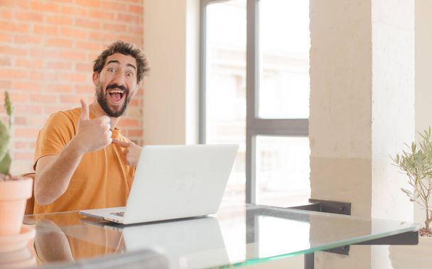 young bearded man feeling proud, carefree, confident and happy, smiling positively with thumbs up and working with a laptop - Foto, Bild