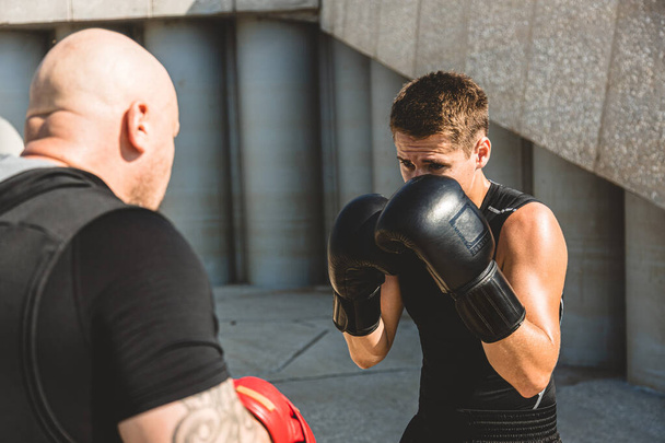 Twee mannen die buiten trainen en vechten. Boxer in handschoenen traint met een coach - Foto, afbeelding