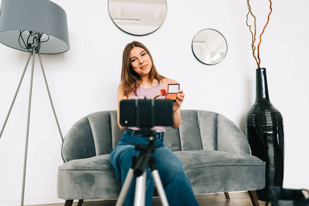 Attractive caucasian young woman video blogger showing beauty products via her blog on social media, holding makeup brushes and shadows looking at smartphone camera fixed on tripod. - Foto, Imagen