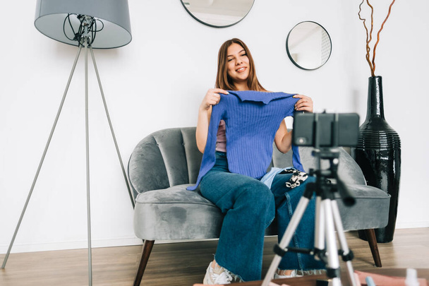 Attractive caucasian young woman video blogger showing new clothing via her blog on social media, holding blouse and looking at smartphone camera fixed on tripod. - Foto, imagen