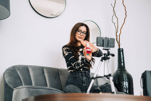 Attractive caucasian young woman video blogger showing beauty products via her blog on social media, looking at smartphone camera fixed on tripod - Photo, Image