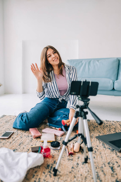Attractive caucasian young woman video blogger showing beauty products via her blog on social media, holding makeup brushes looking at smartphone camera fixed on tripod - Foto, imagen