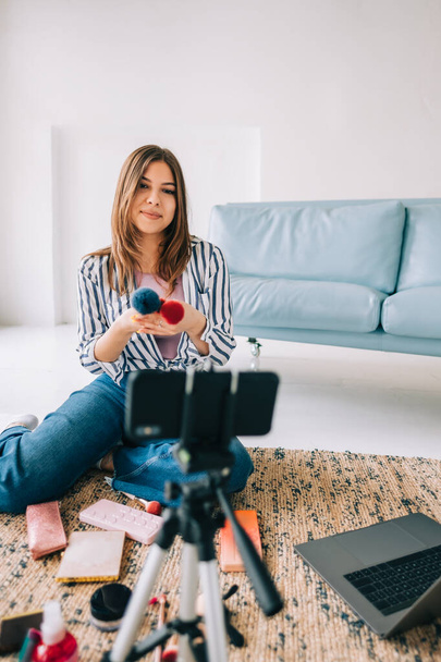 Attractive caucasian young woman video blogger showing beauty products via her blog on social media, holding makeup brushes looking at smartphone camera fixed on tripod - Foto, Imagen