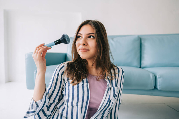 Portrait of attractive caucasian young woman holding makeup brush in hand sitting on floor near blue sofa. - Foto, immagini