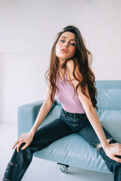 Attractive stylish caucasian woman with long brunette hair posing on blue sofa in modern living room with white walls. - Φωτογραφία, εικόνα