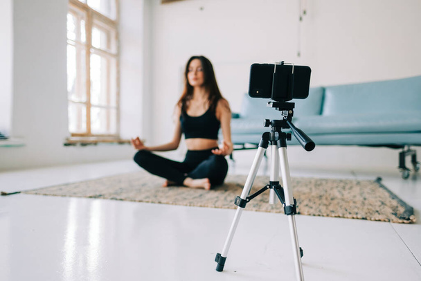 Beautiful brunette fitness woman meditate in front camera on tripod, doing yoga at home. Staying fit and health - Photo, image