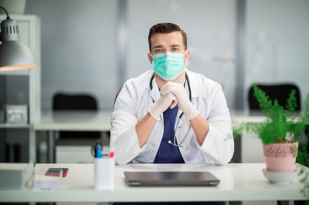 Ein junger Therapeut mit medizinischer Maske sitzt in seinem Büro in einer Privatklinik - Foto, Bild