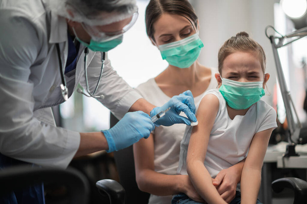 Pieni tyttö pelkää neulan näkemistä ja kipua rokotuksen aikana lääketieteellisessä laboratoriossa, hänen äitinsä tukee häntä. - Valokuva, kuva