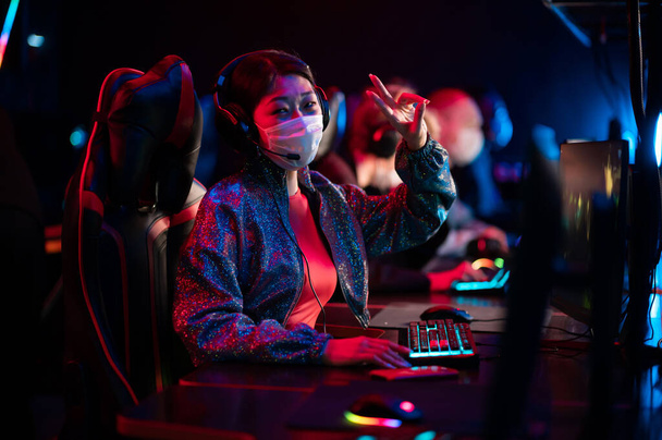 Cute asian woman in a medical mask poses for a photo during a dota game, shows a victory gesture - Фото, изображение
