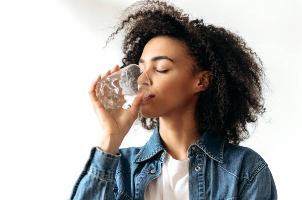 Een gezonde levensstijl. Afro-Amerikaanse krullende jonge vrouw drinkt met plezier een glas zuiver water. Mooie gezonde vrouw volgen gezonde levensstijl, geeft om gezondheid - Foto, afbeelding