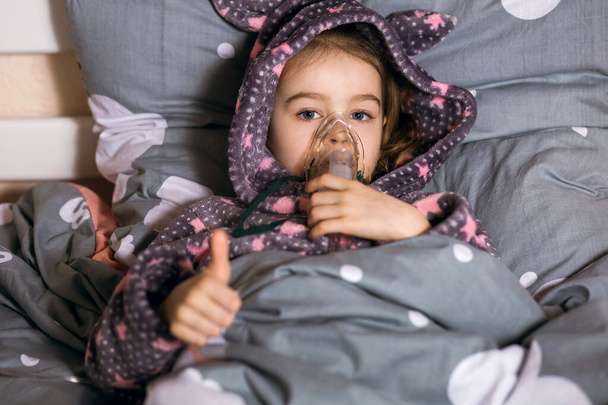 A little girl is being treated for a cough at home using an inhaler. The child is wearing a robe with a hood and the girl shows a thumb up gesture. Optimistic little girl being treated by steam - Photo, Image