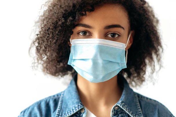 Close-up of african-american curly brunette girl, stands indoors with a protective medical mask on her face and looks directly into the camera. Quarantine precautions, healthcare concept - Photo, Image