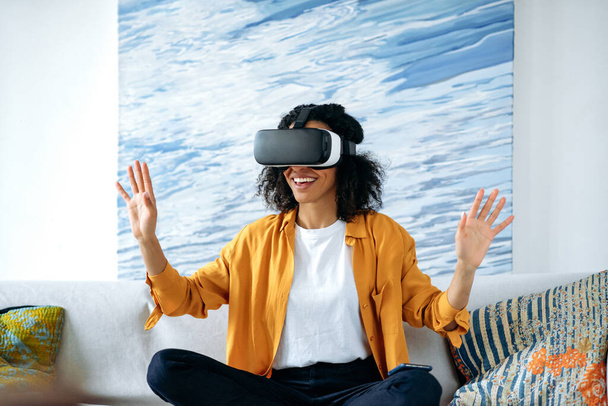 Emocionada chica afroamericana despreocupada, sentada en un sofá en casa y haciendo gestos de manos mientras prueba la nueva aplicación a través del dispositivo VR. Alegre rizado chica moderna con gafas VR, concepto de realidad virtual - Foto, Imagen