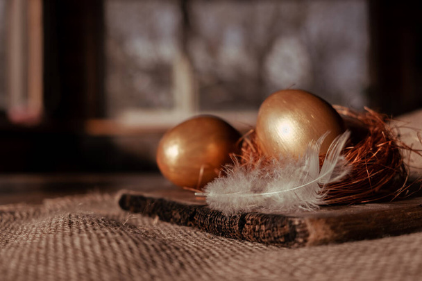 golden eggs lie with nest of straw on canvas cloth against background of window. next to it is a delicate white feather - Photo, image