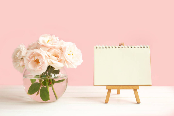 Tiernas rosas rosadas de jardín en un jarrón transparente y un caballete con papel de cuaderno en blanco para escribir deseos. - Foto, imagen