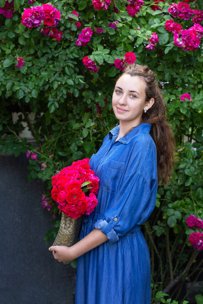 Hermosa adolescente en vestido de mezclilla con flores en el bolso en el jardín de rosas. - Foto, Imagen