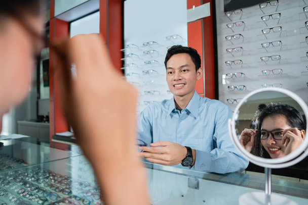 a woman wearing glasses and reflecting in a glass against the background of an eyeglass window display and an employee - Foto, Imagem