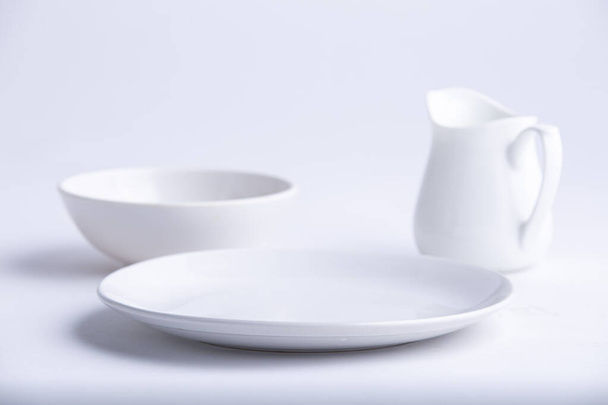  Articles de table blancs, différentes formes d'une plaque en céramique blanche vide, bol et théière disposés sur un fond texturé blanc. - Photo, image