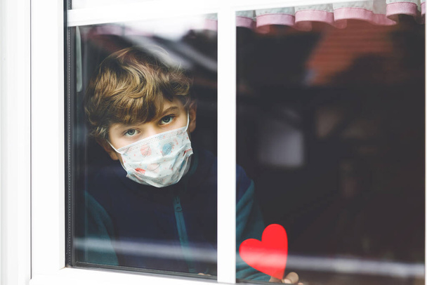 Lovely little school kid boy by a window wearing medical mask and holding wooden heart during pandemic coronavirus quarantine. Lonely upset child, self isolation concept. - Photo, Image