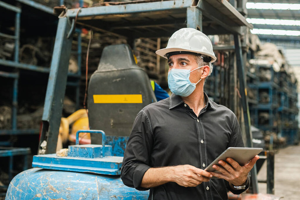 人の工学の労働者は保護面マスクの均一および倉庫で働くためのタブレットを保持するヘルメットを身に着けている。職場工場産業用重機. - 写真・画像