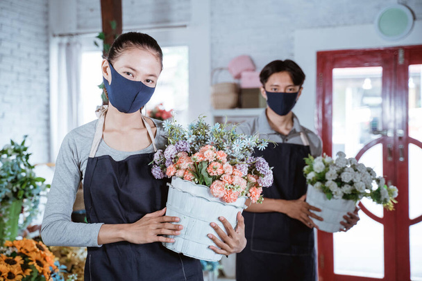 young female florist wearing apron and face mask holding bucket flower smiling looking at camera. working in flower shop - Fotografie, Obrázek