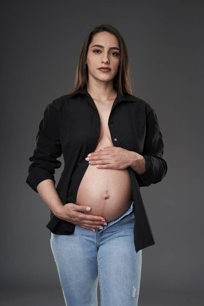 Pregnant woman in blue jeans and unbuttoned black shirt posing on gray background - Φωτογραφία, εικόνα