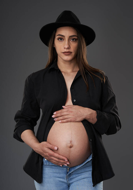 Pregnant woman in blue jeans, hat and unbuttoned black shirt posing on gray background - Foto, Imagen