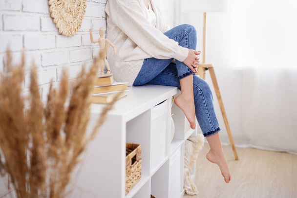 home and cosiness concept - young slim woman sitting on shelf in living room - 写真・画像