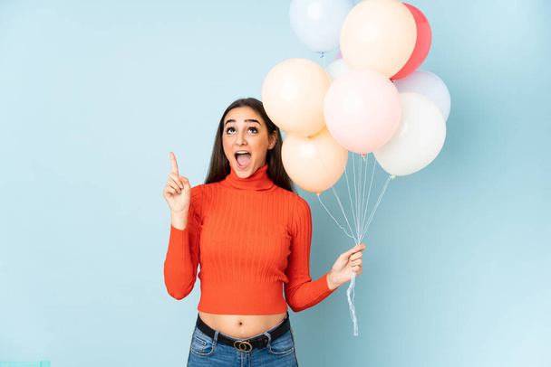 Young woman catching many balloons isolated on blue background pointing with the index finger a great idea - Fotografie, Obrázek
