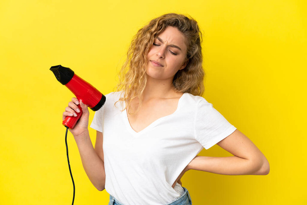 Young blonde woman holding a hairdryer isolated on yellow background suffering from backache for having made an effort - Photo, Image