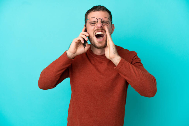 Jonge Braziliaanse man met behulp van mobiele telefoon geïsoleerd op blauwe achtergrond schreeuwen met de mond wijd open - Foto, afbeelding