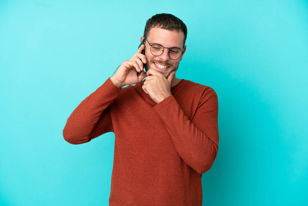 携帯電話を使っている若いブラジル人男性側を見て笑っている青い背景に隔離された - 写真・画像