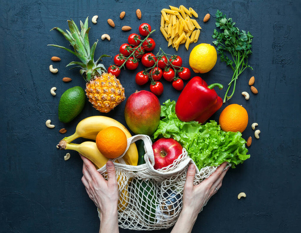 Hands holding a string bag with healthy vegetarian food. Variety of vegetables and fruits on a black background. Food shopping, zero waste and clean vegan eating concept. - 写真・画像