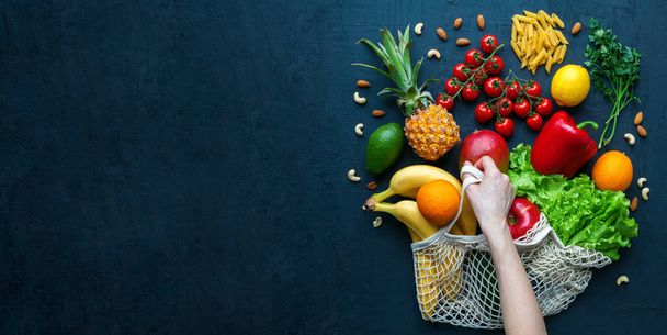 Human hand holding a string bag with healthy vegetarian food. Variety of vegetables and fruits on a black background, copy-space, banner. Food shopping, zero waste and clean vegan eating concept. - Photo, image