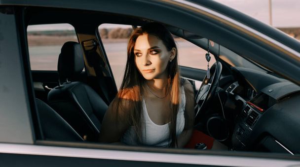 Beautiful young girl driving a car in the evening in the sunset sun on an empty parking lot - Foto, afbeelding