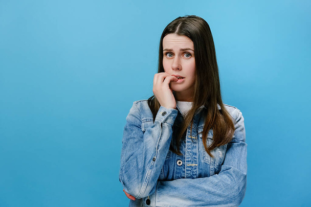 Worried young girl folded hands together near mouth, gnawing nails, young woman feels uncertain, stressed. Confused brunette female with frighten or panic gesture, isolated on blue studio background - Foto, Imagem