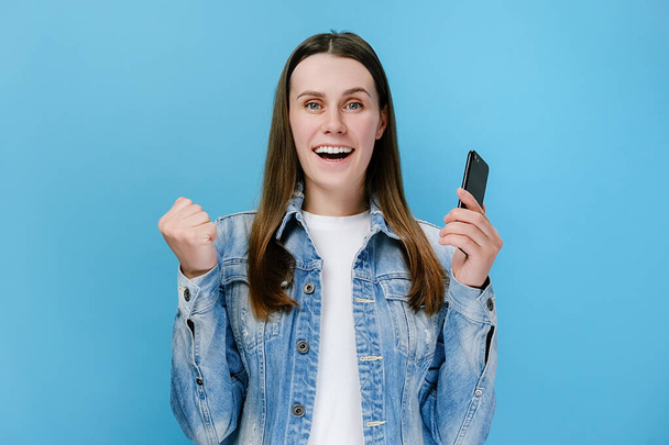 Excited cheerful young woman holds smartphone receive sms with great news feels happy make yes gesture, wears denim jacket, isolated on blue background. Moment of victory showing sincere emotions - Photo, image