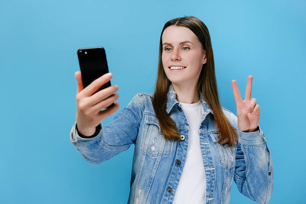 Lovely playful young woman blogger taking selfie with mobile phone, makes peace gesture, has fun with cellular, shooting social media stories, wears denim jacket, poses against blue studio background - Photo, image