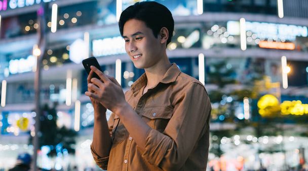 Молодой азиат пользуется телефоном, гуляя по улице ночью. - Фото, изображение
