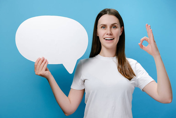 Portrait of cute beautiful young happy woman holding speech bubble posing isolated over blue studio background wall with copy space make okay gesture, dressed in white t-shirt. Hand sign concept - Photo, Image