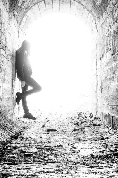 Μια νεαρή γυναίκα στην άκρη του τούνελ. Το κορίτσι στέκεται κοντά στον τοίχο στις ακτίνες του φωτός. Ασπρόμαυρη φωτογραφία με κοκκώδες αποτέλεσμα - Φωτογραφία, εικόνα