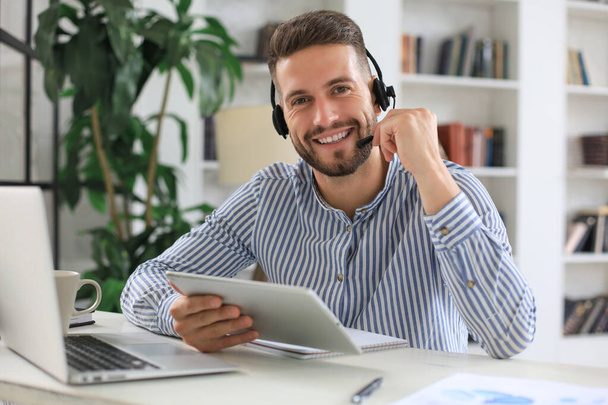 Cheerful young support phone male operator in headset, at workplace while using digital tablet, help service and client consulting call center concept. - Fotó, kép